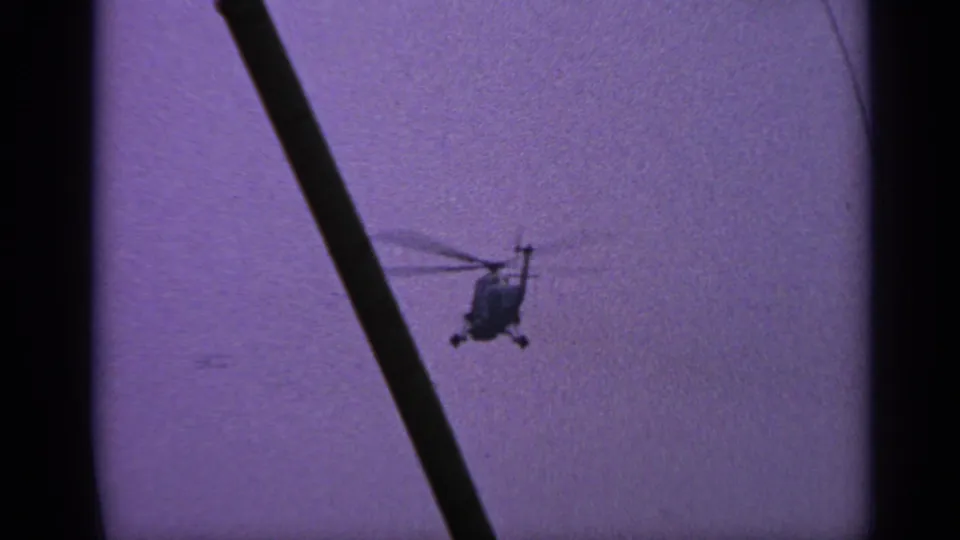 1970s United Kingdom home-movie frame showing the helicopter farther left and partially obstructed by a dark foreground pole.
