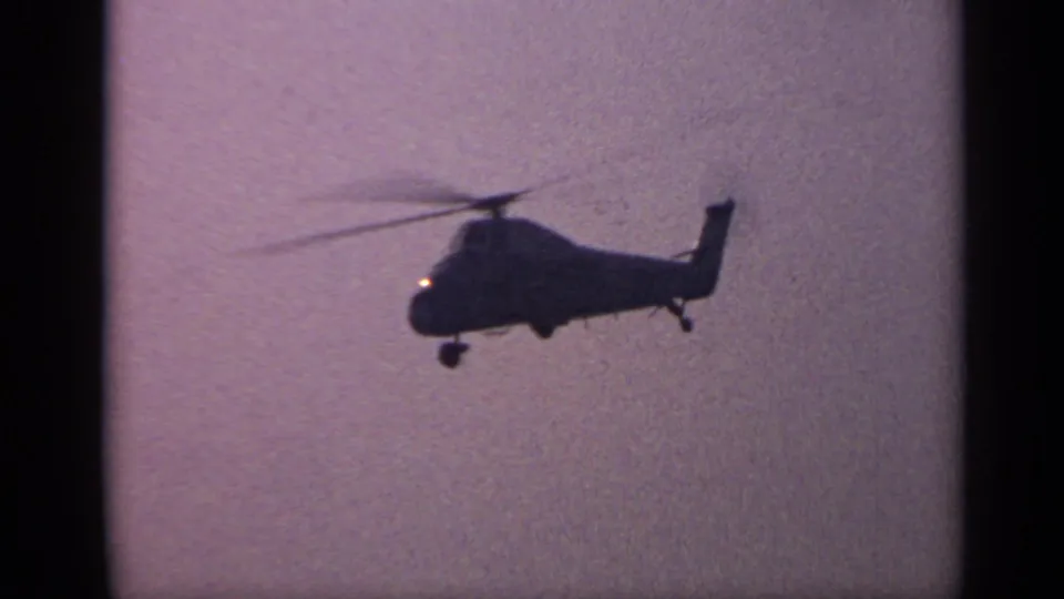 1970s United Kingdom 8mm frame showing a closer helicopter silhouette, with landing gear and rotor blur visible against a tinted sky.