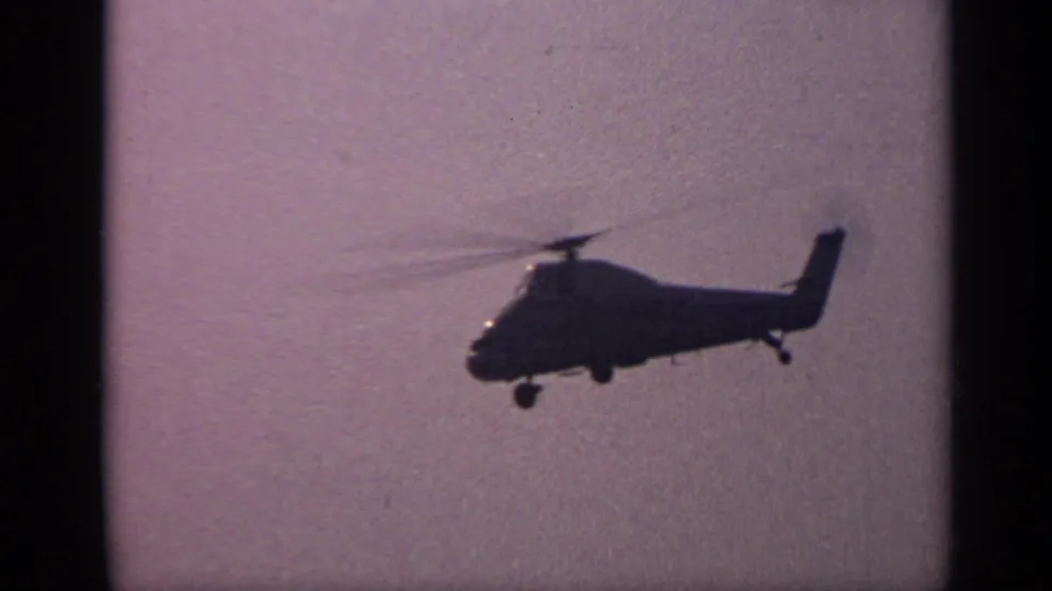 1970 United Kingdom home-movie frame of a helicopter profile in flight, seen as a dark silhouette against a soft purple sky.