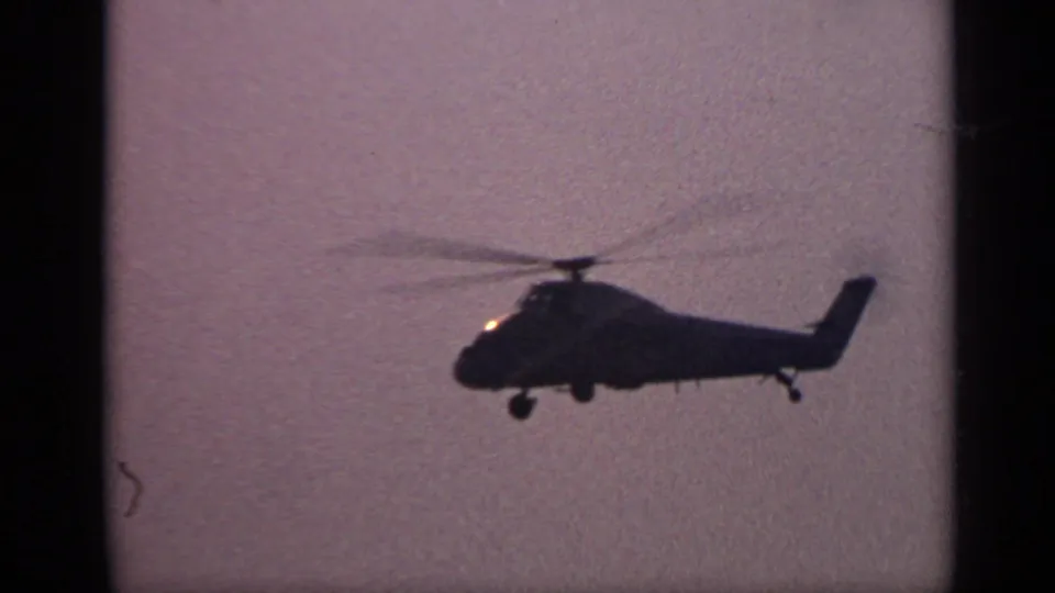 A large helicopter in silhouette flies over water in 1970 United Kingdom 8mm home-movie footage.