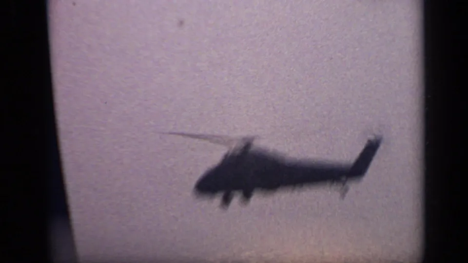 A blurred large helicopter crosses low over water, with motion and handheld focus visible in the 8mm frame.