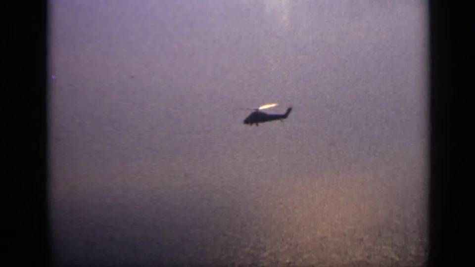 The helicopter appears smaller and farther away above reflective water in the final 1970 United Kingdom frame.