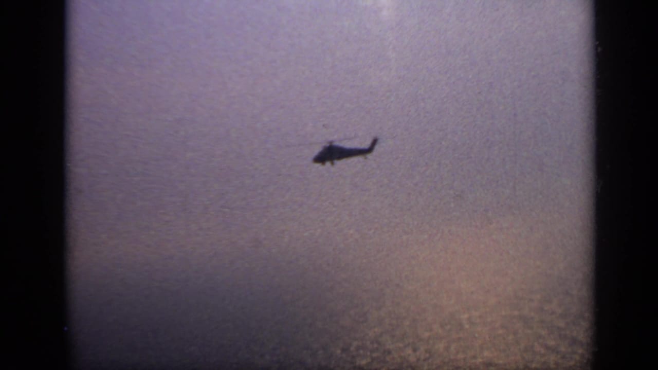 Helicopter seen in silhouette over a broad body of water, with 1970s 8mm grain and subdued color.