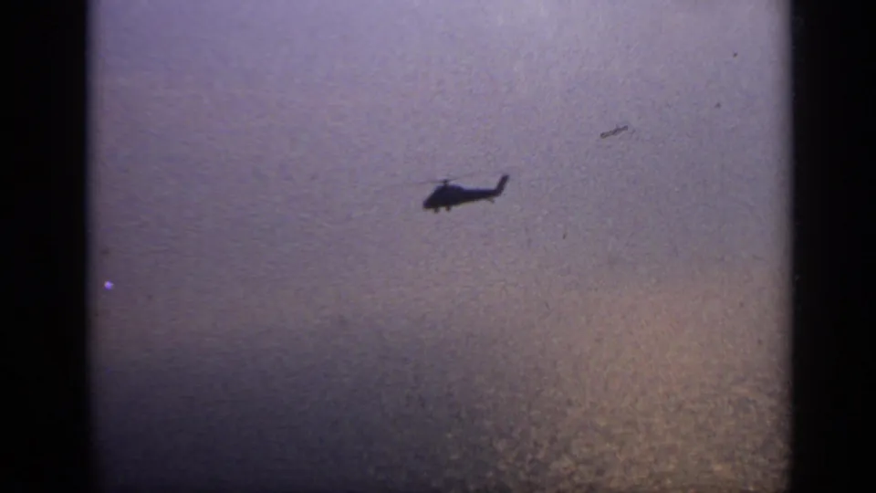 A small helicopter silhouette is filmed from afar over textured water in archival 8mm footage.