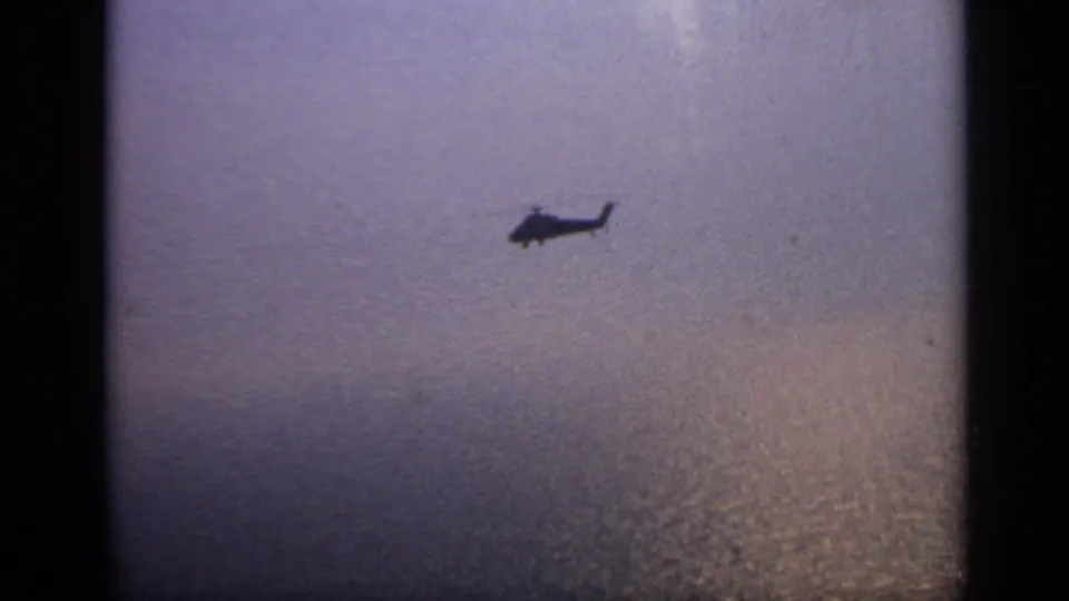 Distant helicopter silhouette over water, framed by grainy 8mm film and soft evening-like color.