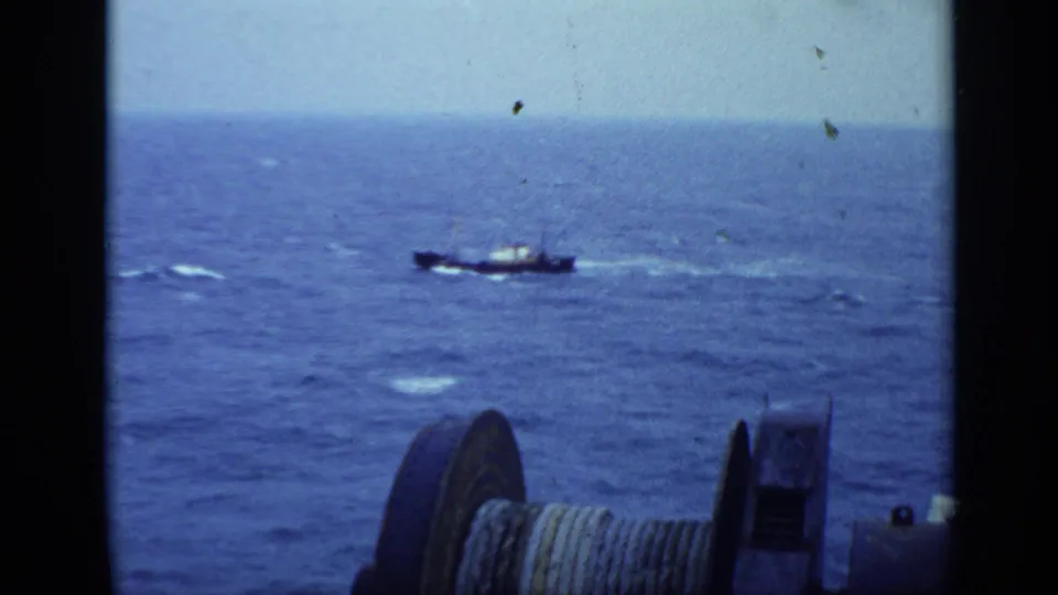 A small vessel moves across open water, viewed from another ship with a large cable spool in the foreground.