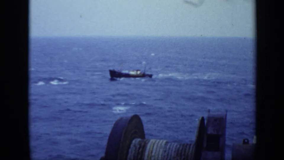 The fishing vessel remains distant on choppy water, with a large shipboard cable spool visible at the lower frame edge.