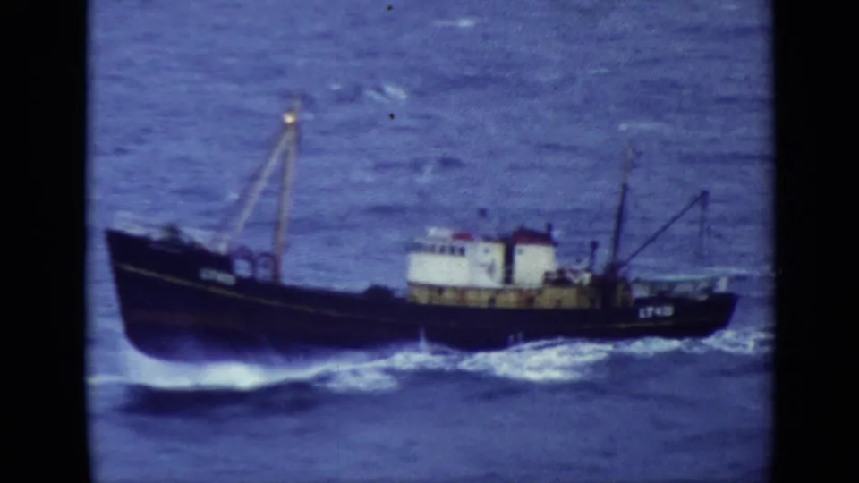 A dark fishing vessel is seen closer in frame, pushing through rough blue water with white spray along the hull.