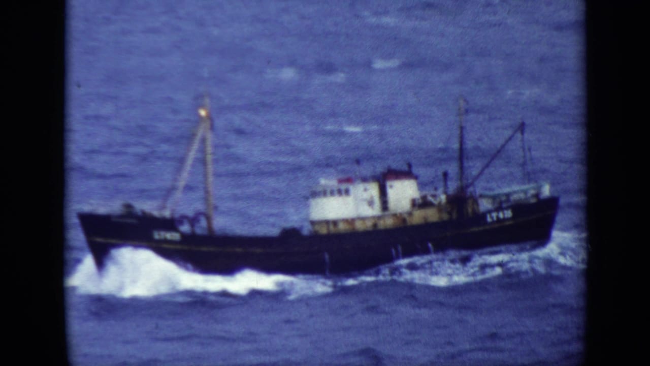 Dark fishing trawler moving across choppy blue water with white spray at the bow.
