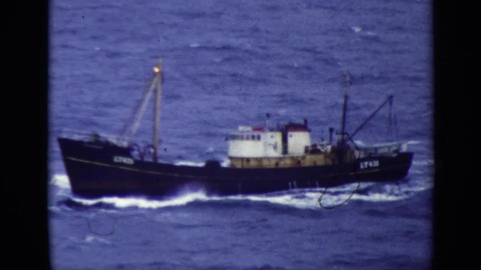1970 United Kingdom 8mm frame of a dark-hulled fishing trawler moving across choppy blue water with rigging and a white wake.