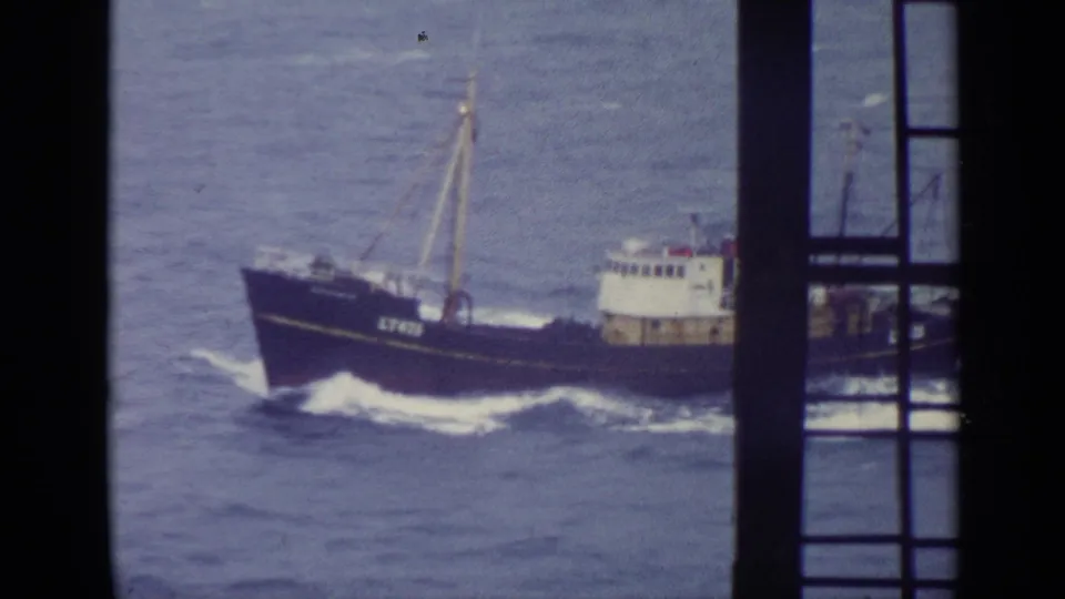 The dark fishing trawler continues across choppy sea, partly blocked by a vertical window frame in 1970 United Kingdom home-movie footage.