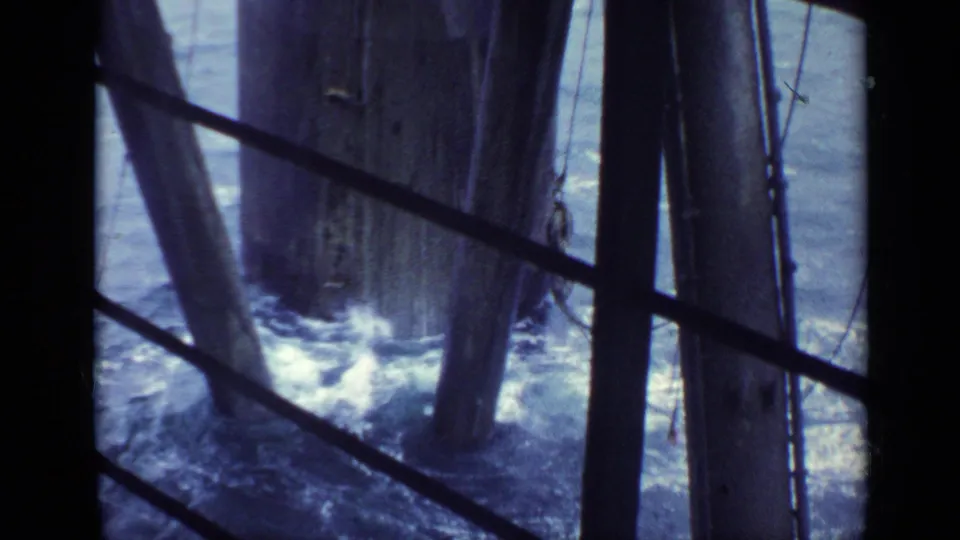 Choppy water splashes around heavy vertical supports, viewed through dark railings or window structure in the same 1970 United Kingdom 8mm clip.