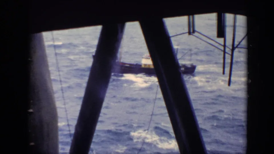 The small brown boat passes behind heavy structural beams, with choppy sea and whitecaps filling the frame.