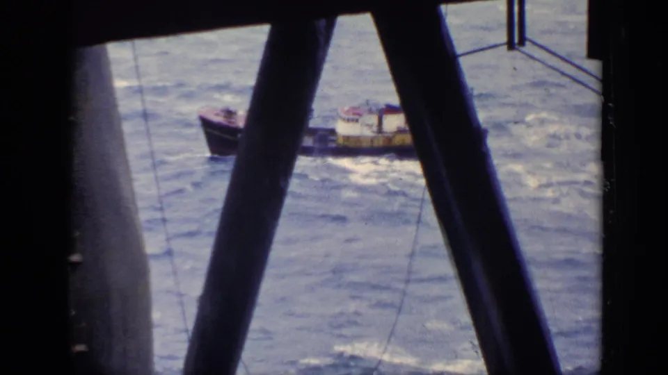 A brown boat rides over choppy water near a larger structure, framed by dark beams in archival 8mm footage.