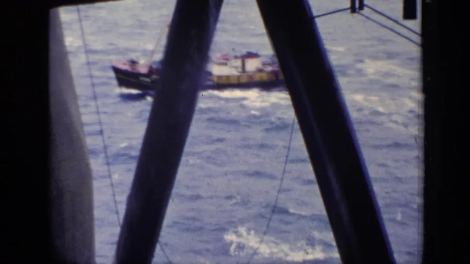 1970 United Kingdom 8mm frame showing a brown tugboat on choppy water, viewed through dark offshore or vessel structure.