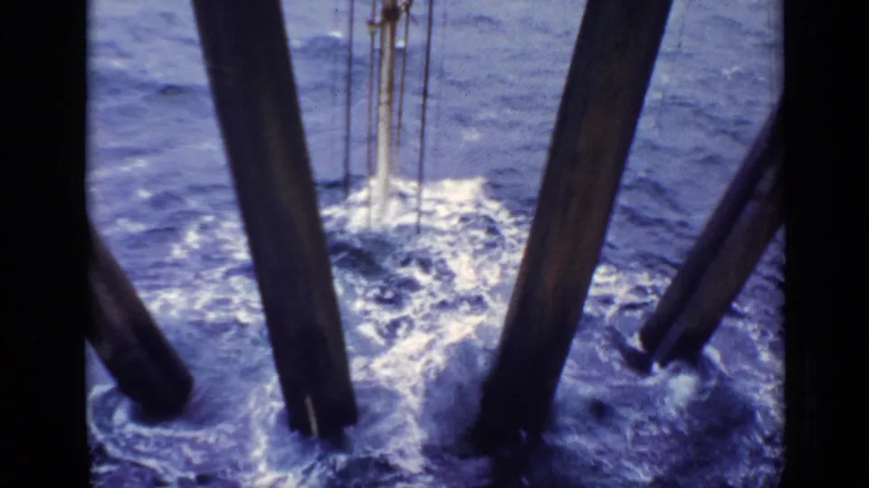 1970 United Kingdom 8mm frame of rough sea and white foam seen through dark beams from an elevated maritime structure.
