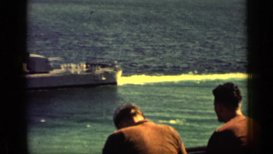 Two men on shore or a dock watch the water as the rear of a naval ship exits the 1962 Cuba home-movie frame.