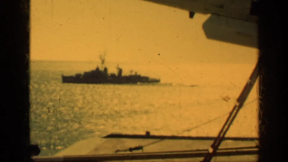 1962 Cuba 8mm frame of a distant naval-looking vessel at sea, seen over railings and deck hardware from another ship.