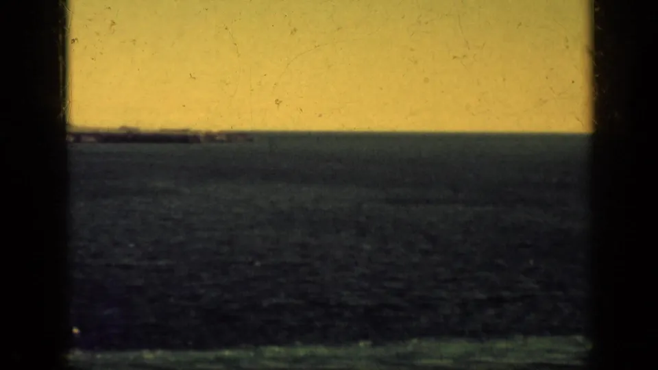 Yellow-tinted 8mm view of open water and a faint horizon line from a shipboard window, with no clear landmarks visible.