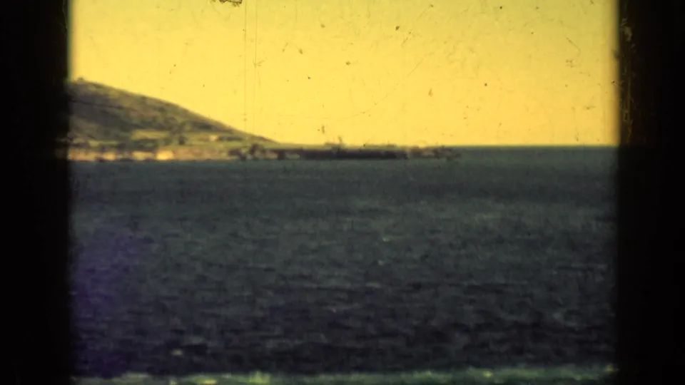 1962 Cuba 8mm frame of a coastal hillside across the water, with a dark window frame and warm degraded color.