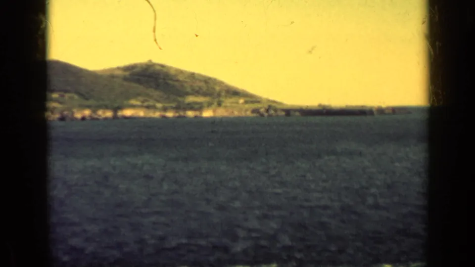 1962 Cuba 8mm view of coastal mountains, shoreline structures, open sea, and a distant dark ship under a yellow sky.