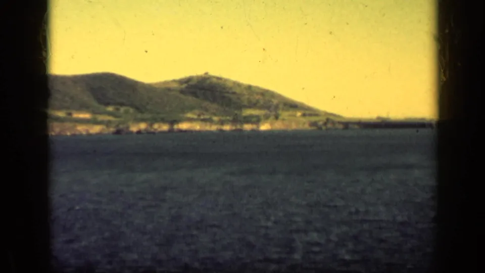 1962 Cuba home-movie frame showing rolling coastal mountains beyond blue water, with dark window framing and a warm yellow sky cast.