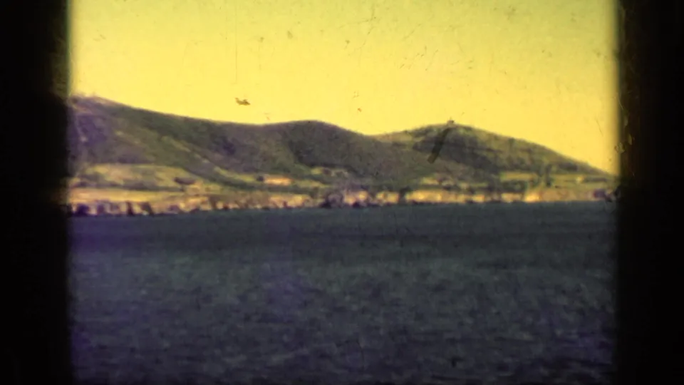 1962 Cuba coastal landscape with a mountain ridge, narrow shoreline, blue water, and heavy dark vertical framing from the filmed viewpoint.