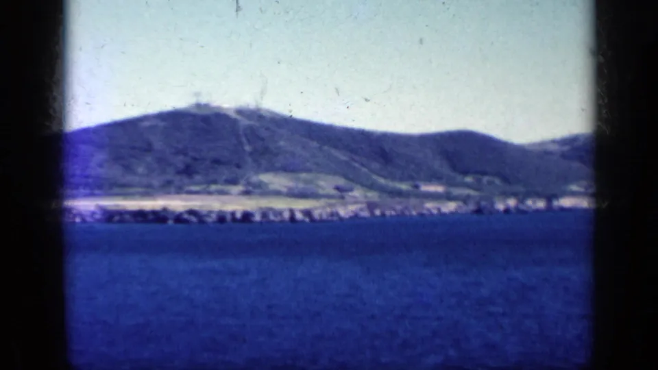 Blue-shifted 1962 Cuba frame showing sea, pale shoreline, and rounded mountains through a dark foreground window or rail division.