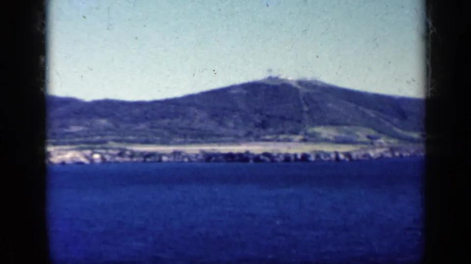 1962 Cuba coastal scenery with a broad blue water foreground, distant shoreline, mountain range, pale sky, and a dark vertical divider in front.