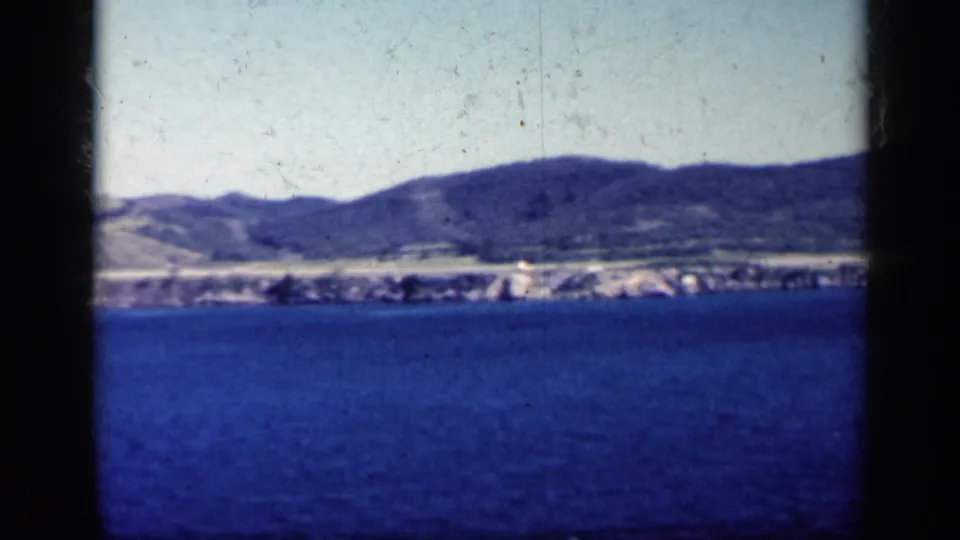 1962 Cuba coastal view from the water, with a low built-up shoreline below rounded hills and a pale sky.