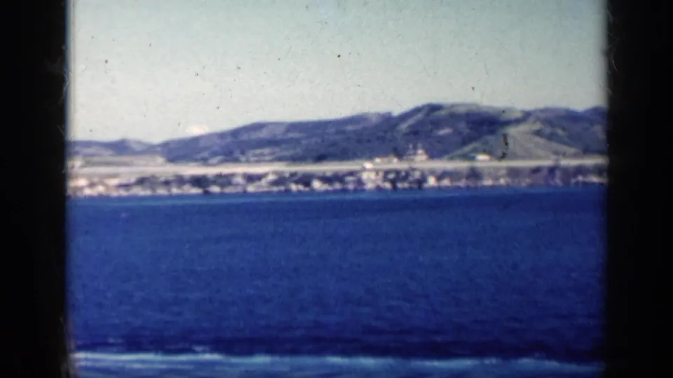 Final 1962 Cuba frame looking over blue water toward a low shoreline and hills, with soft color and dark film borders.