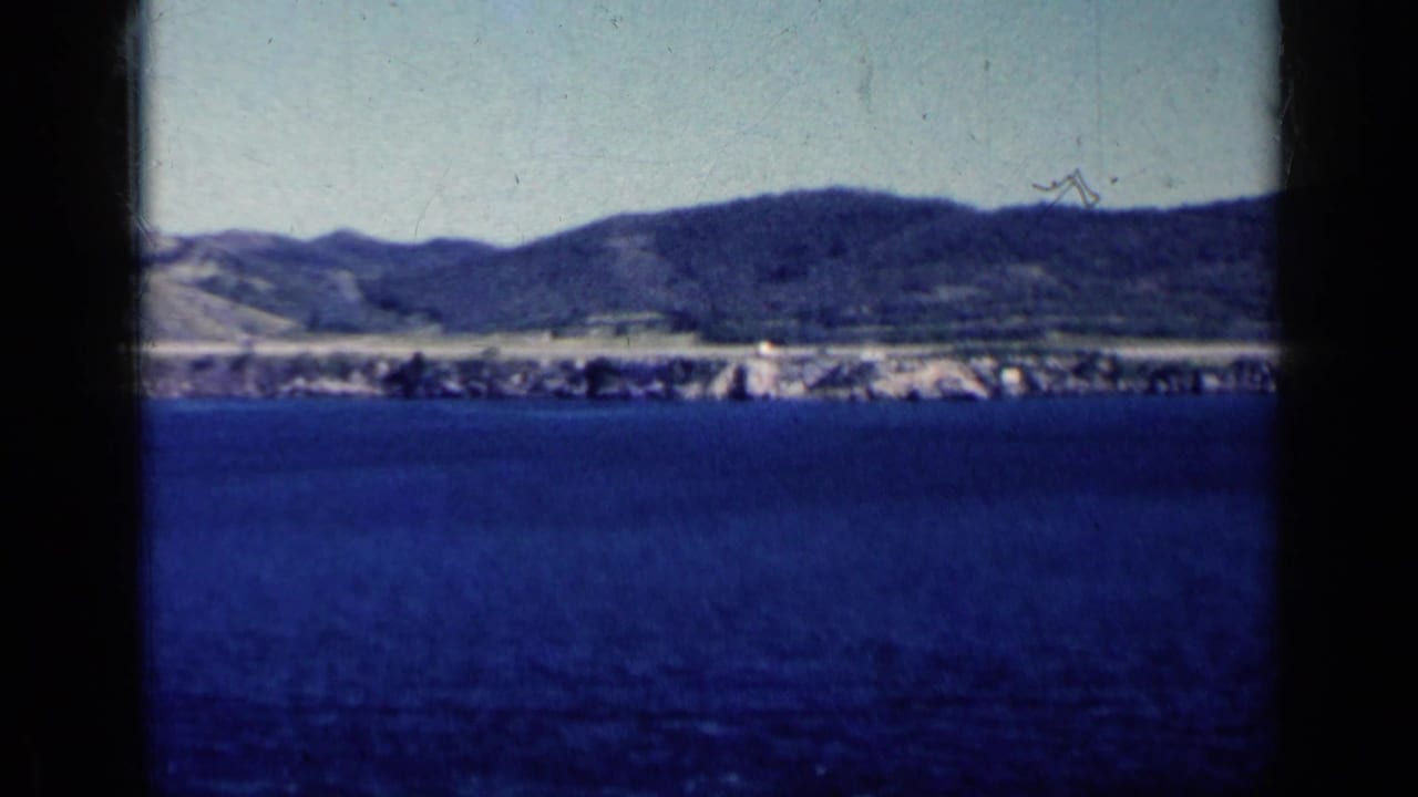 Blue water facing a distant shoreline and hills, framed by dark film or window edges.