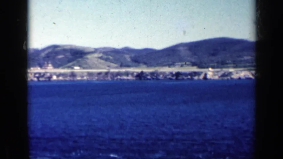 1962 Cuba coastal view with blue water in the foreground, low buildings along shore, and hills beyond.