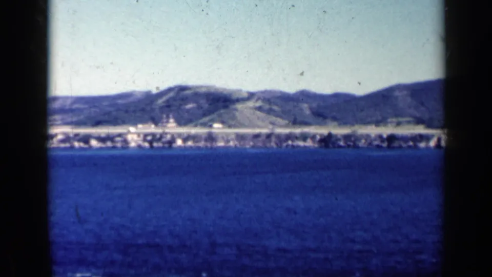 1962 Cuba 8mm frame showing blue water, a low coastal settlement, and rounded hills under a pale sky.