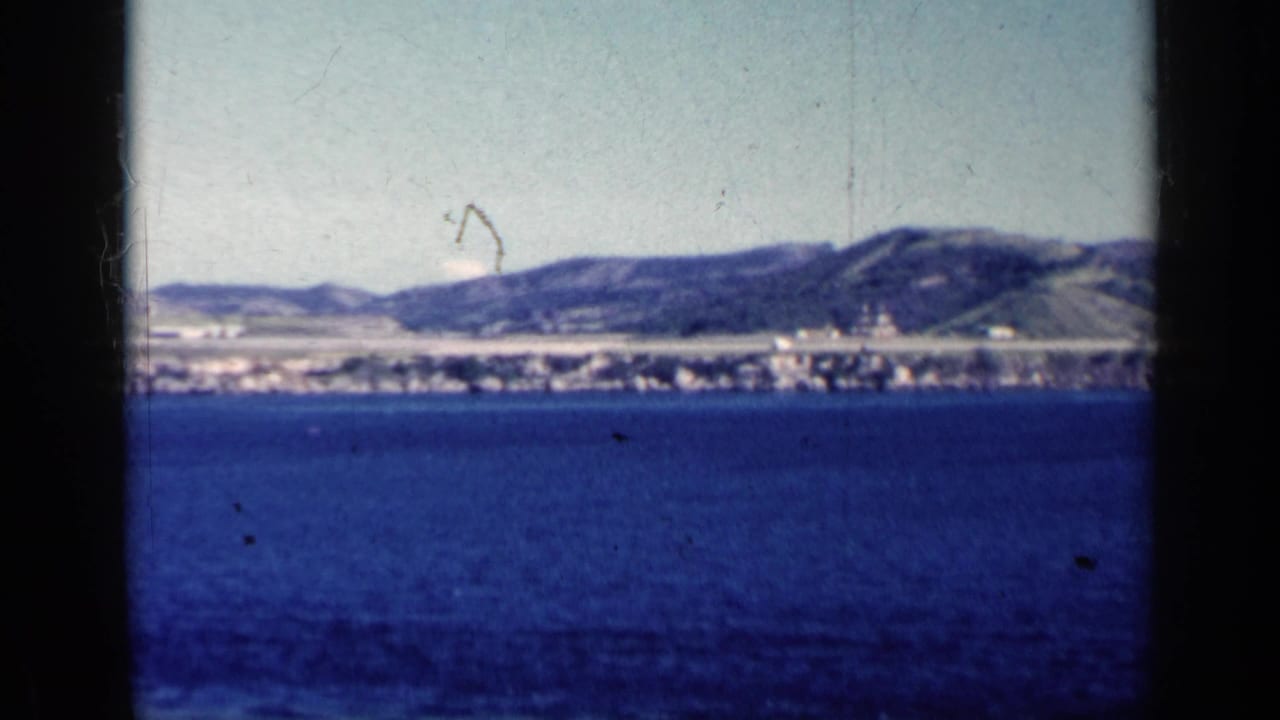 1962 Cuba 8mm frame showing open blue water, a low distant coast, scattered buildings, and hills beyond the shoreline.