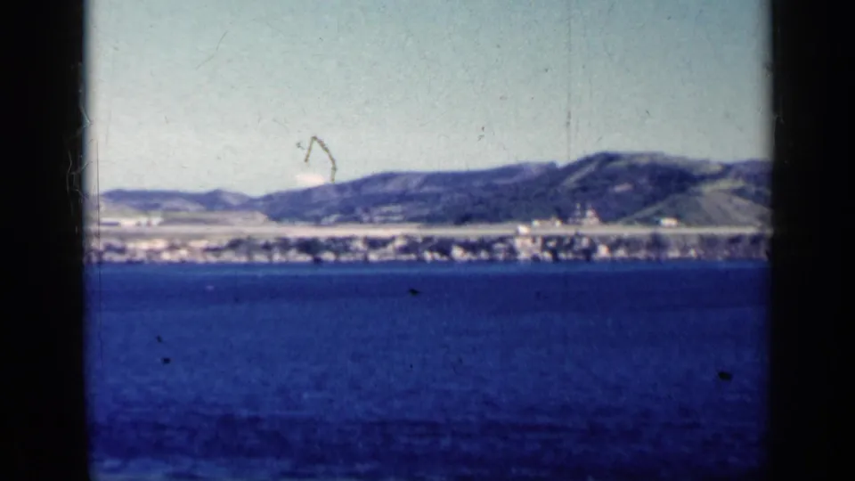 1962 Cuba 8mm frame showing open blue water, a low distant coast, scattered buildings, and hills beyond the shoreline.