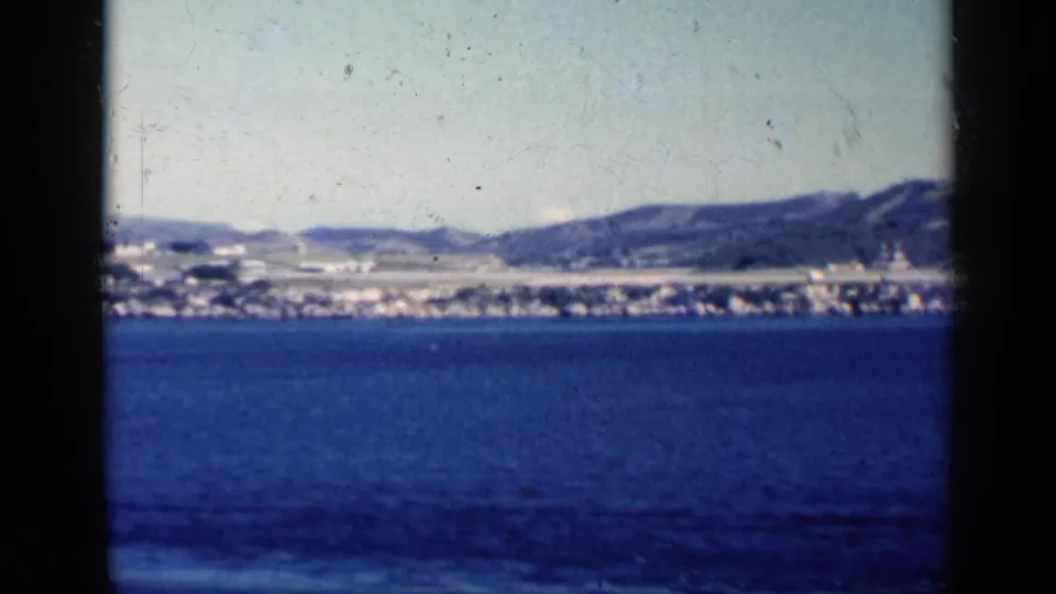 1962 Cuba 8mm view across deep blue water toward a distant coastal strip, low structures, and hills.