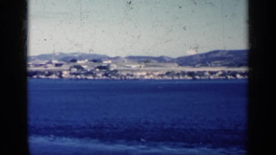 1962 Cuba 8mm frame with ocean in the foreground, a narrow settled shoreline, and hills behind it.