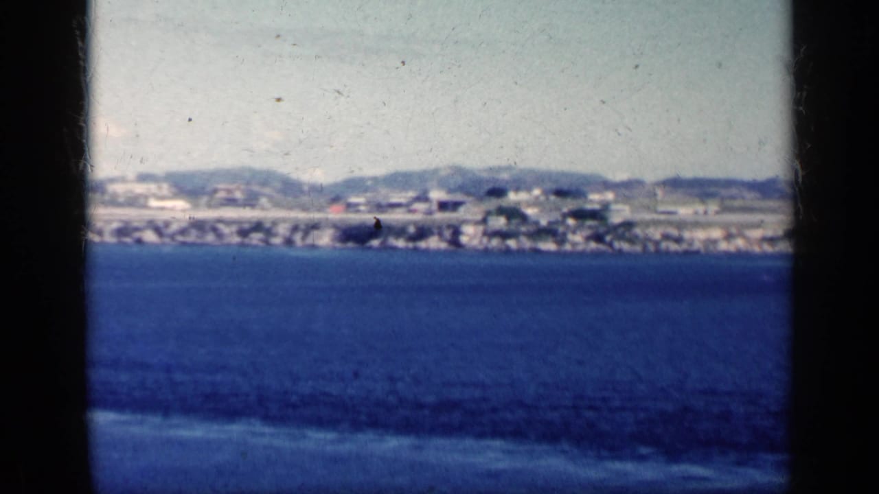 Blue ocean facing a distant shoreline with low buildings and hills under a pale sky.