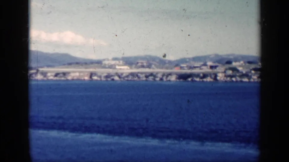 1962 Cuba coastal view across dark blue ocean, with low shoreline buildings, a possible elevated roadway and hazy hills in the background.