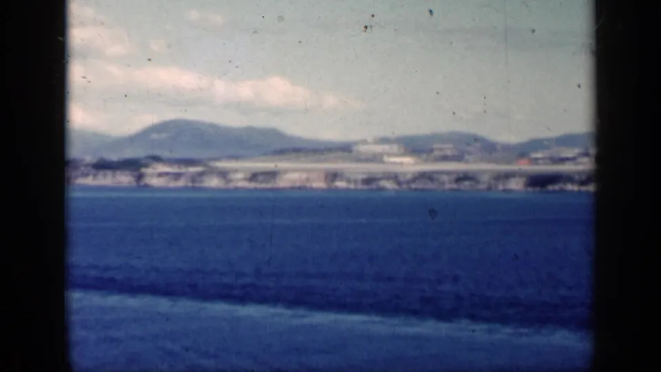 1962 Cuba 8mm frame with blue ocean foreground, distant low-rise waterfront, rolling hills and a cloudy sky in soft home-movie focus.