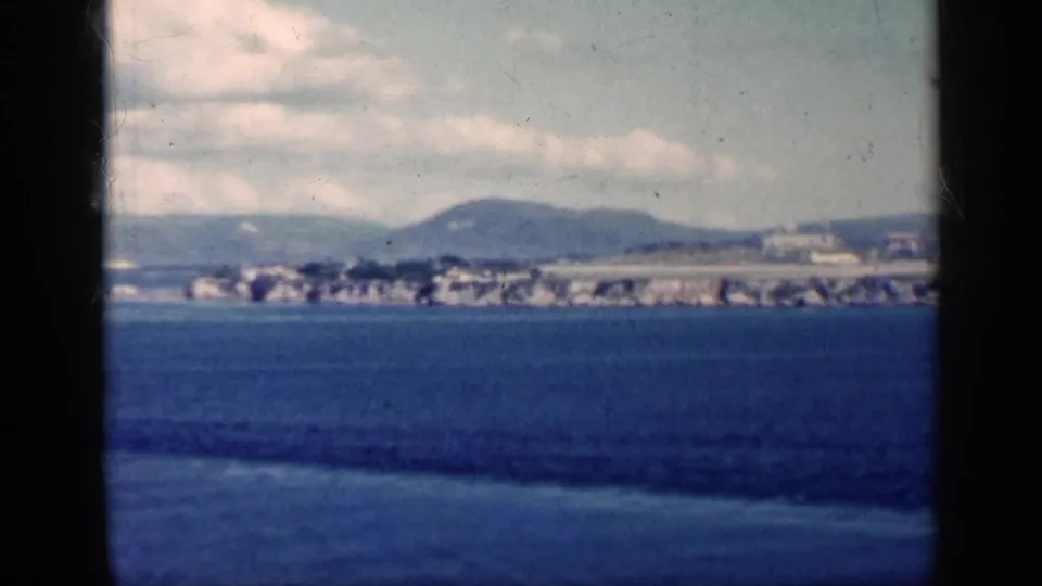 1962 Cuba home-movie frame showing a distant coastal city line across blue water, with hills behind it and scattered clouds above.