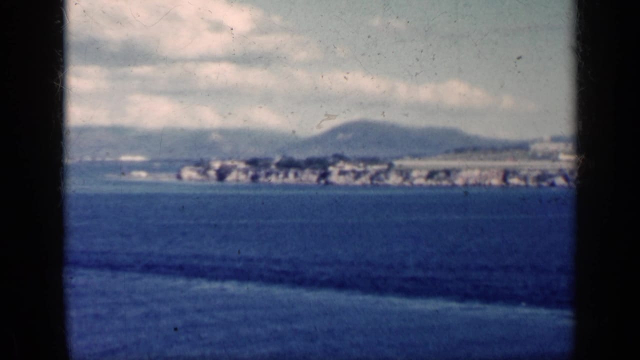 1962 Cuba 8mm frame showing blue water, a distant shoreline with low buildings, hills, and a cloudy sky.