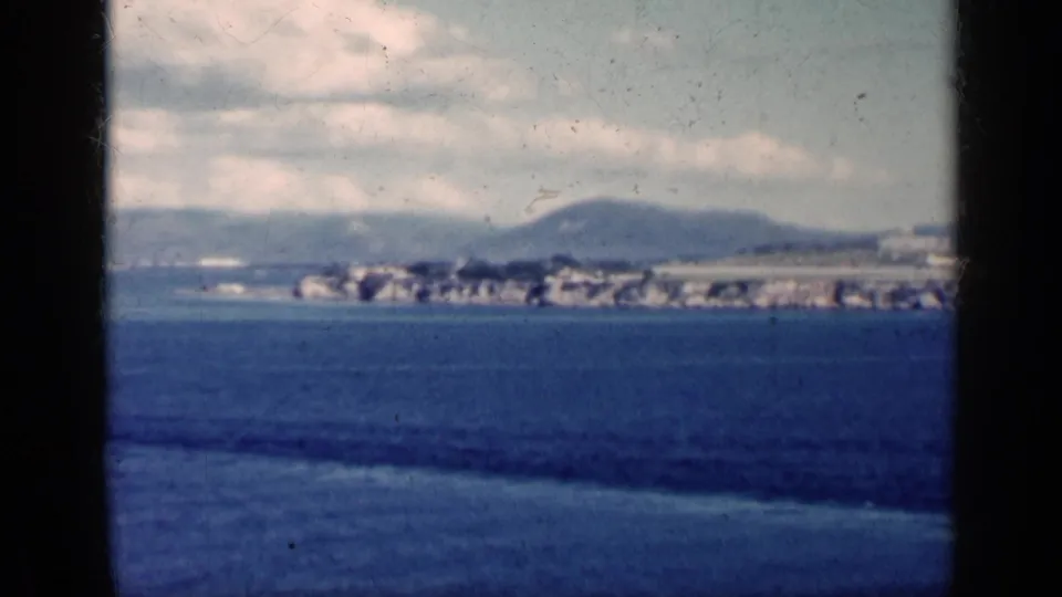 1962 Cuba 8mm frame showing blue water, a distant shoreline with low buildings, hills, and a cloudy sky.