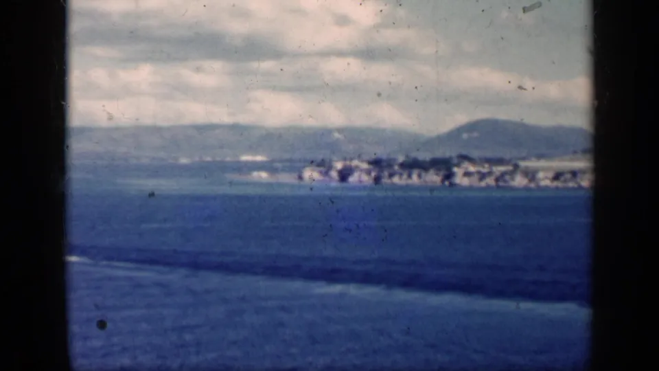 1962 Cuba coastal view across open water, with low waterfront buildings, hills, clouds, and dark frame edges.