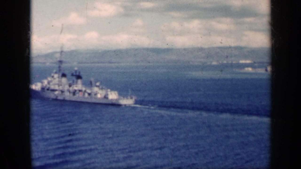 Naval warship crossing blue water with distant coastline under a cloudy sky.
