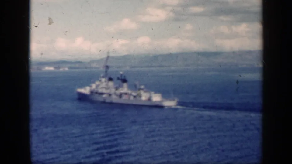 A naval warship appears mid-frame on blue coastal water near Cuba, with soft focus, dark frame borders, and land visible beyond.