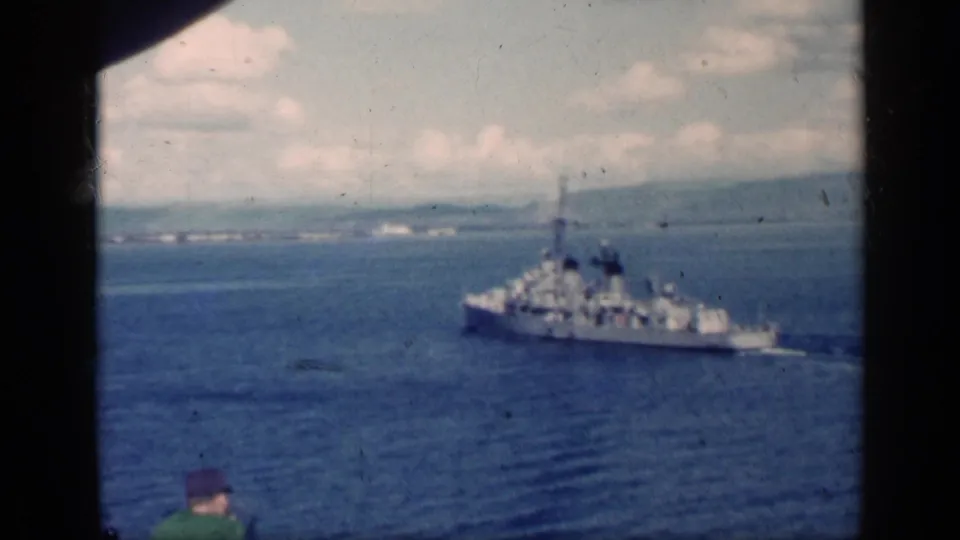 1962 Cuba home-movie frame of a naval warship at sea, with a seated passenger partly visible in the foreground and coastline beyond.