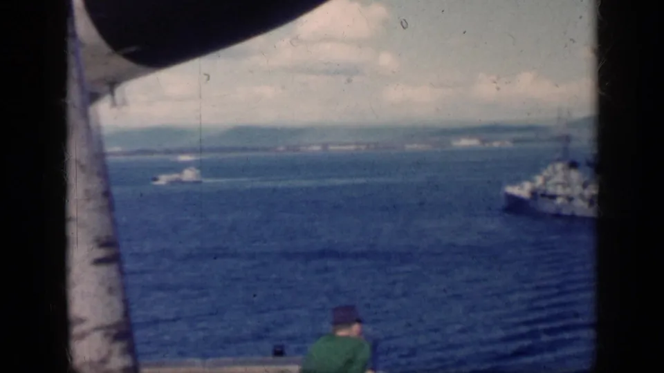 The warship slips partly out of frame at right in 1962 Cuba coastal footage, while a seated passenger and boat structure appear in the foreground.