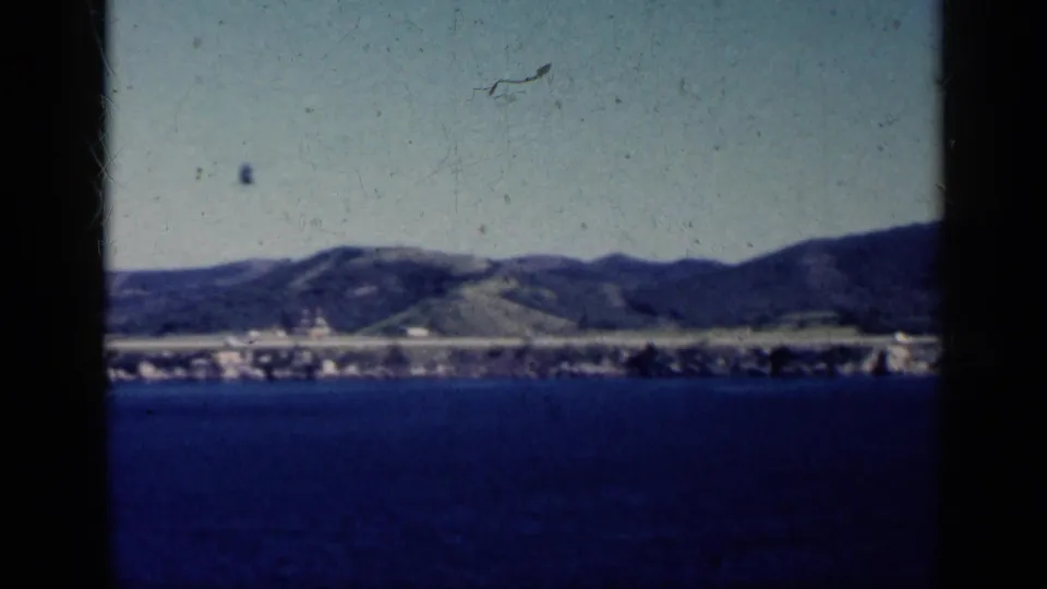 1962 Cuba 8mm frame showing open blue water, distant shoreline structures, and low green hills viewed from offshore.
